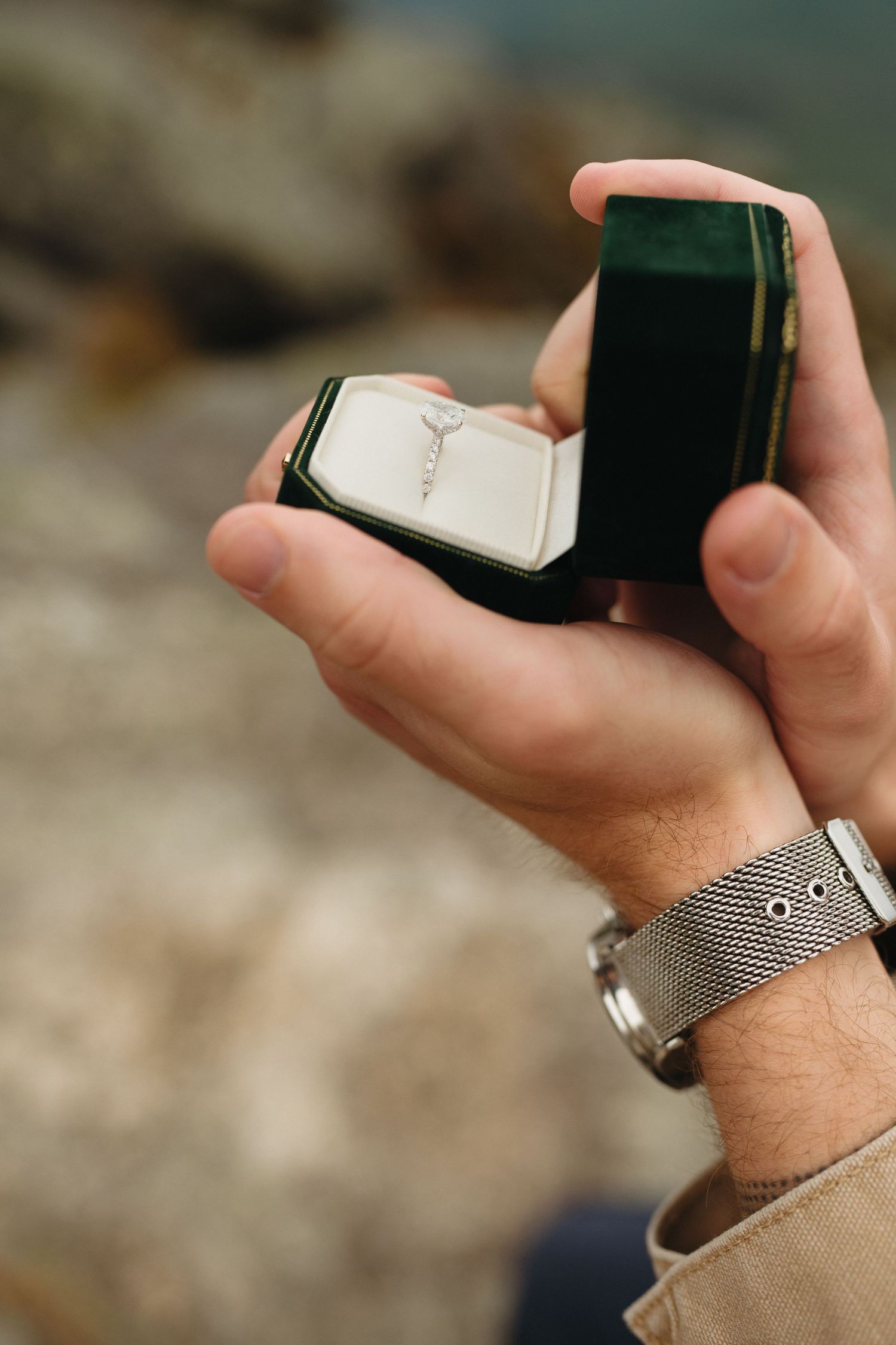 Man holding open green velvet ring box with diamond engagement ring outdoors.