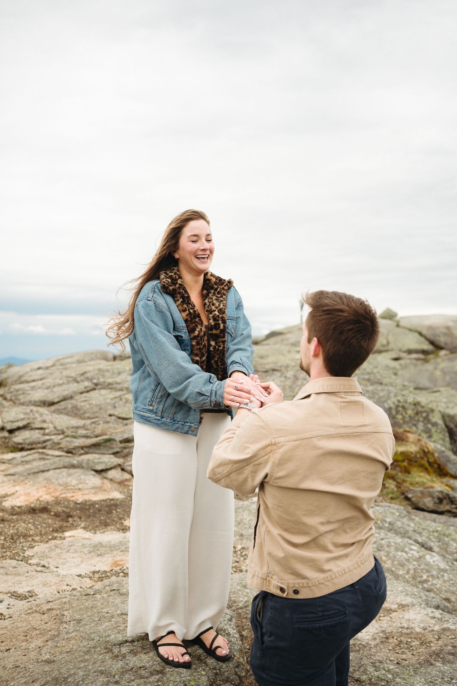 Man kneeling on rocky mountaintop proposing to laughing woman in denim jacket and white pants on cloudy day.
