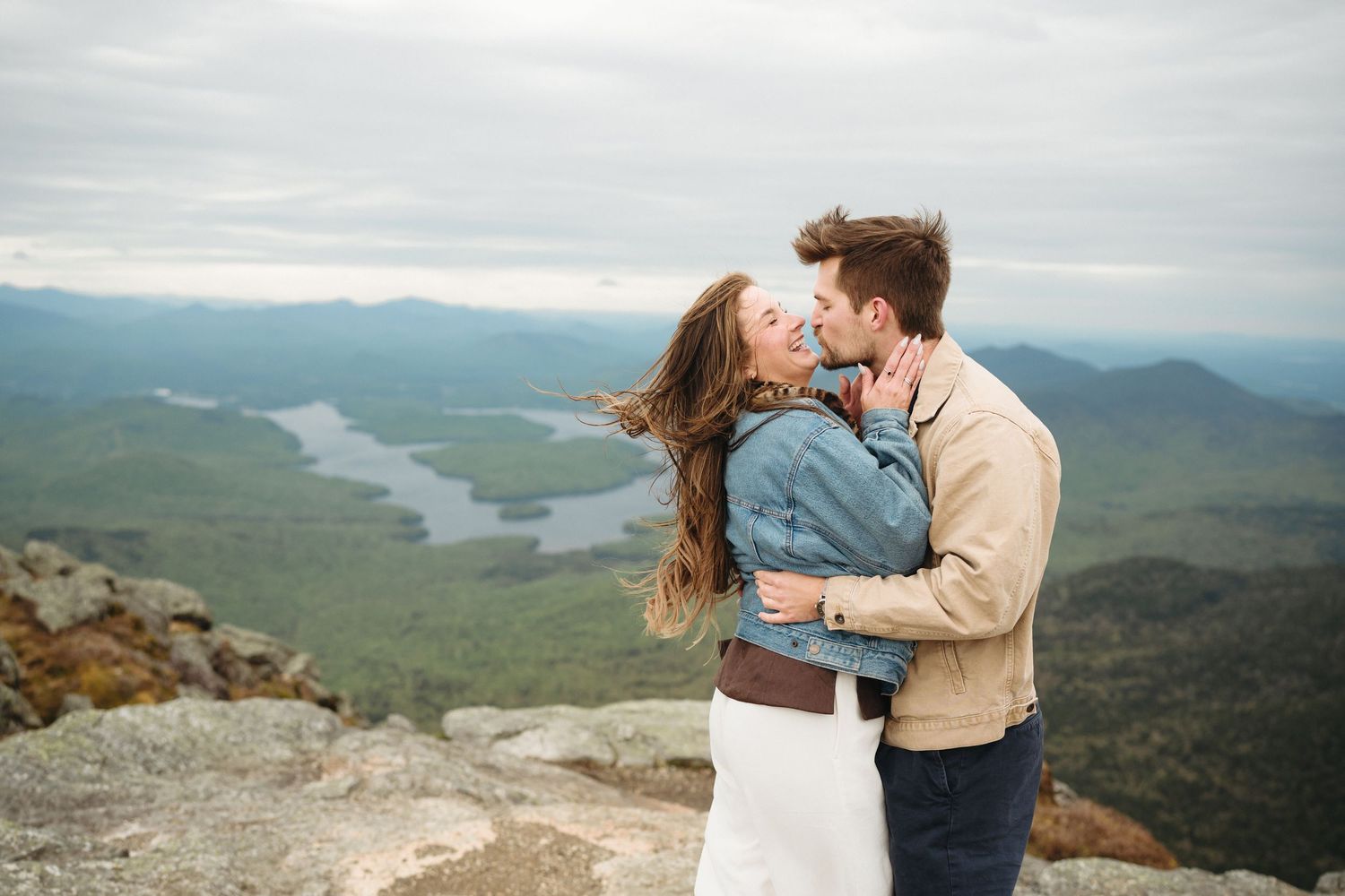 Couple sharing a romantic kiss on a mountain summit with sweeping valley and lake views behind them.