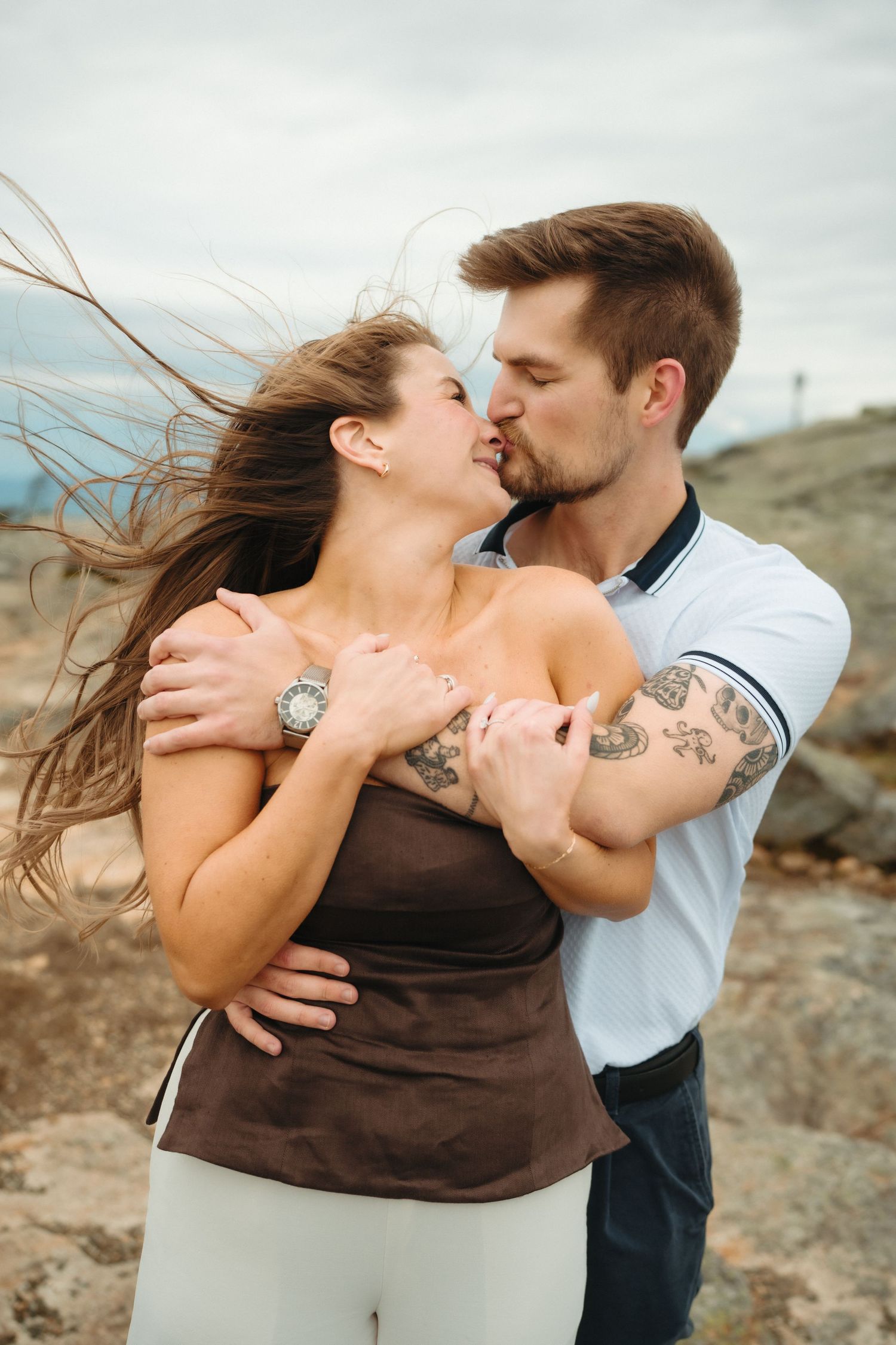 A couple shares a romantic kiss outdoors on rocky terrain, with the woman's hair blowing in the wind.
