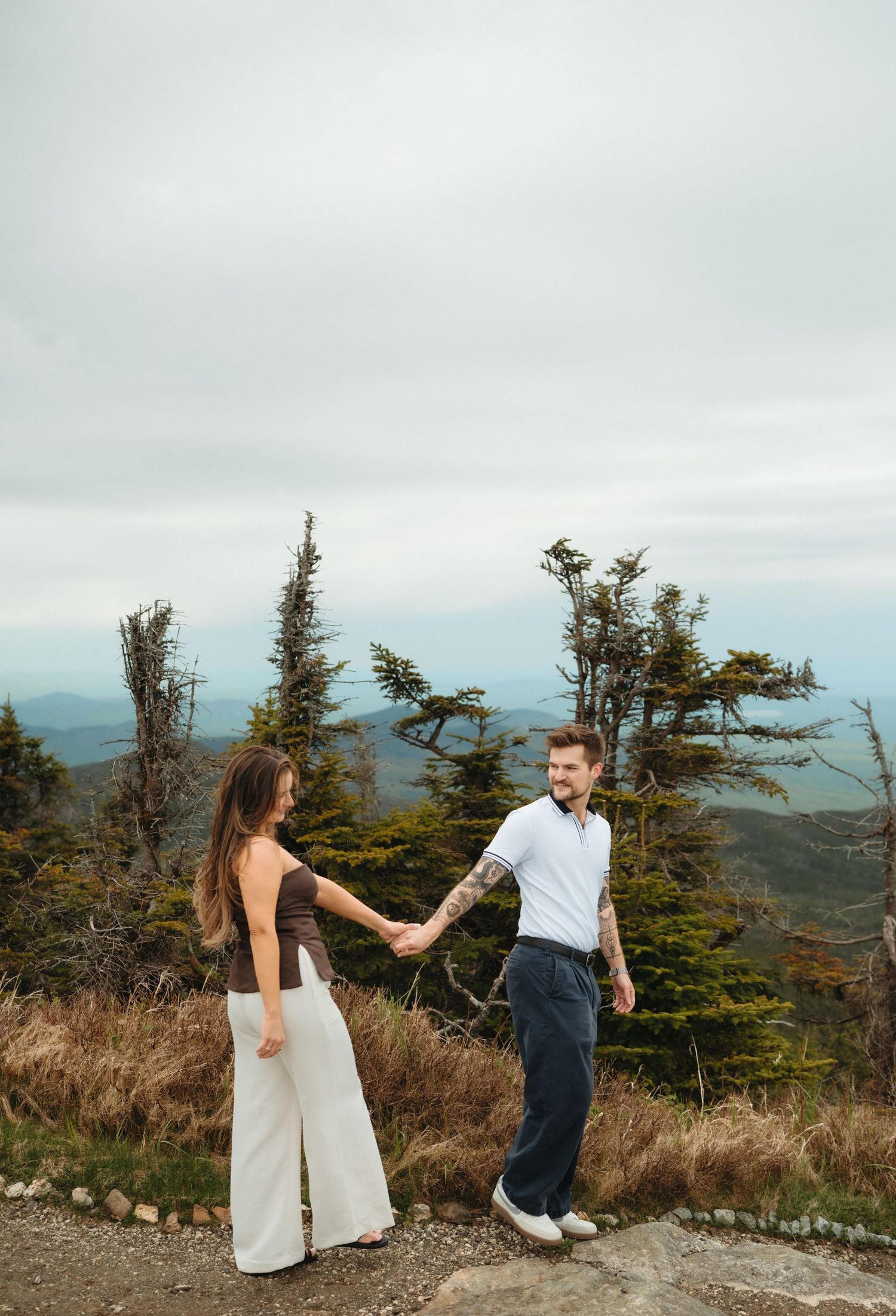 Couple holding hands on rocky mountain summit with scenic valley views.
