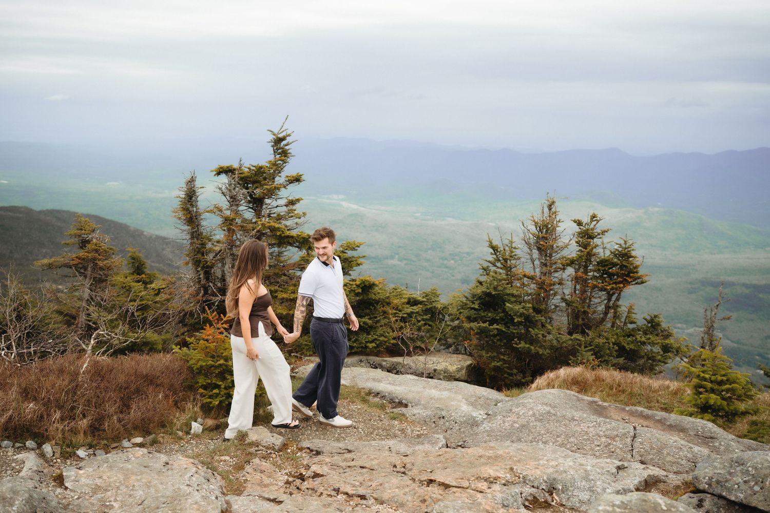 Couple holding hands on rocky mountain summit with lush green valley and misty hills in background.