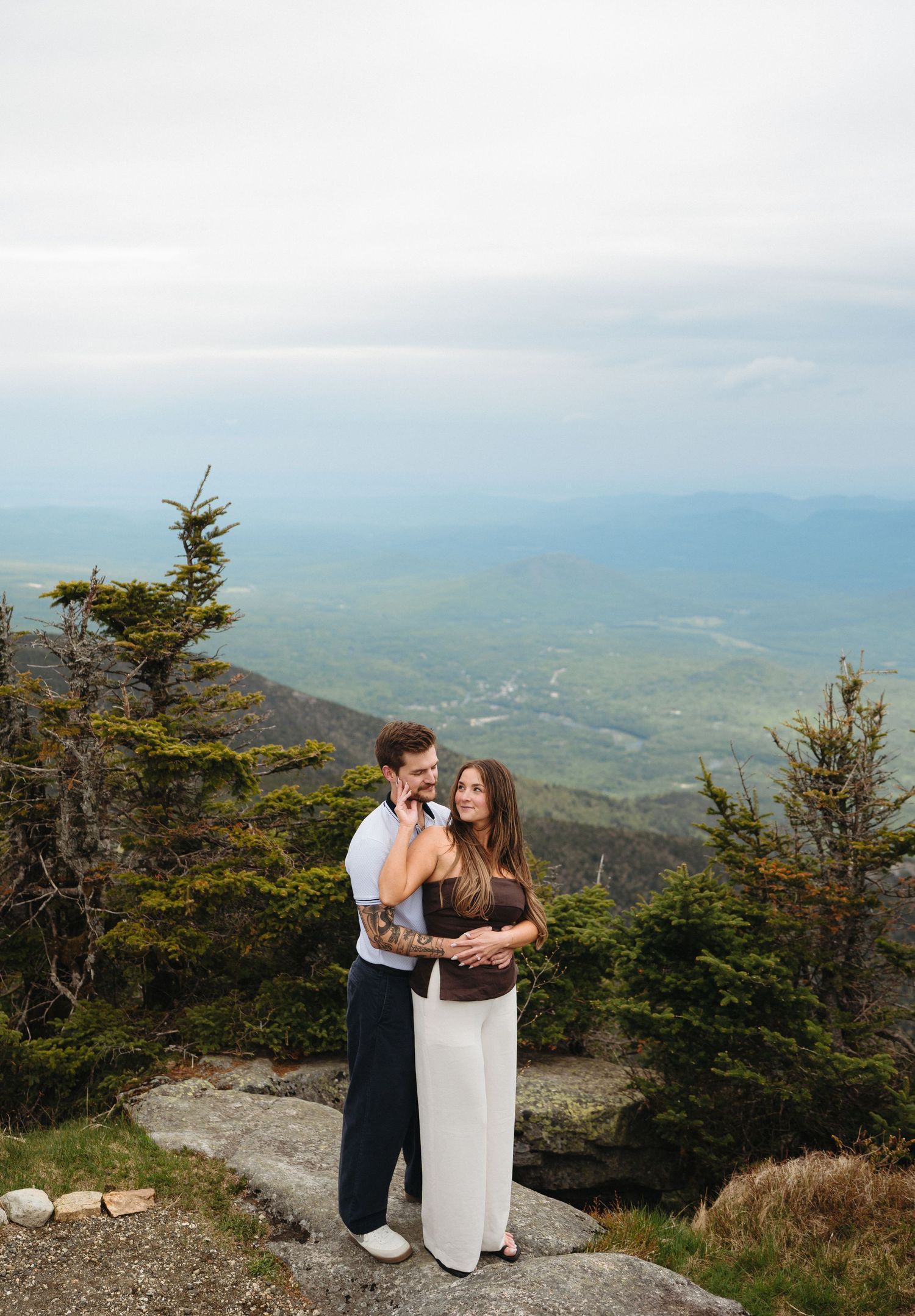 Couple embracing on a rocky mountain summit with sweeping green valley views and evergreen trees behind them.
