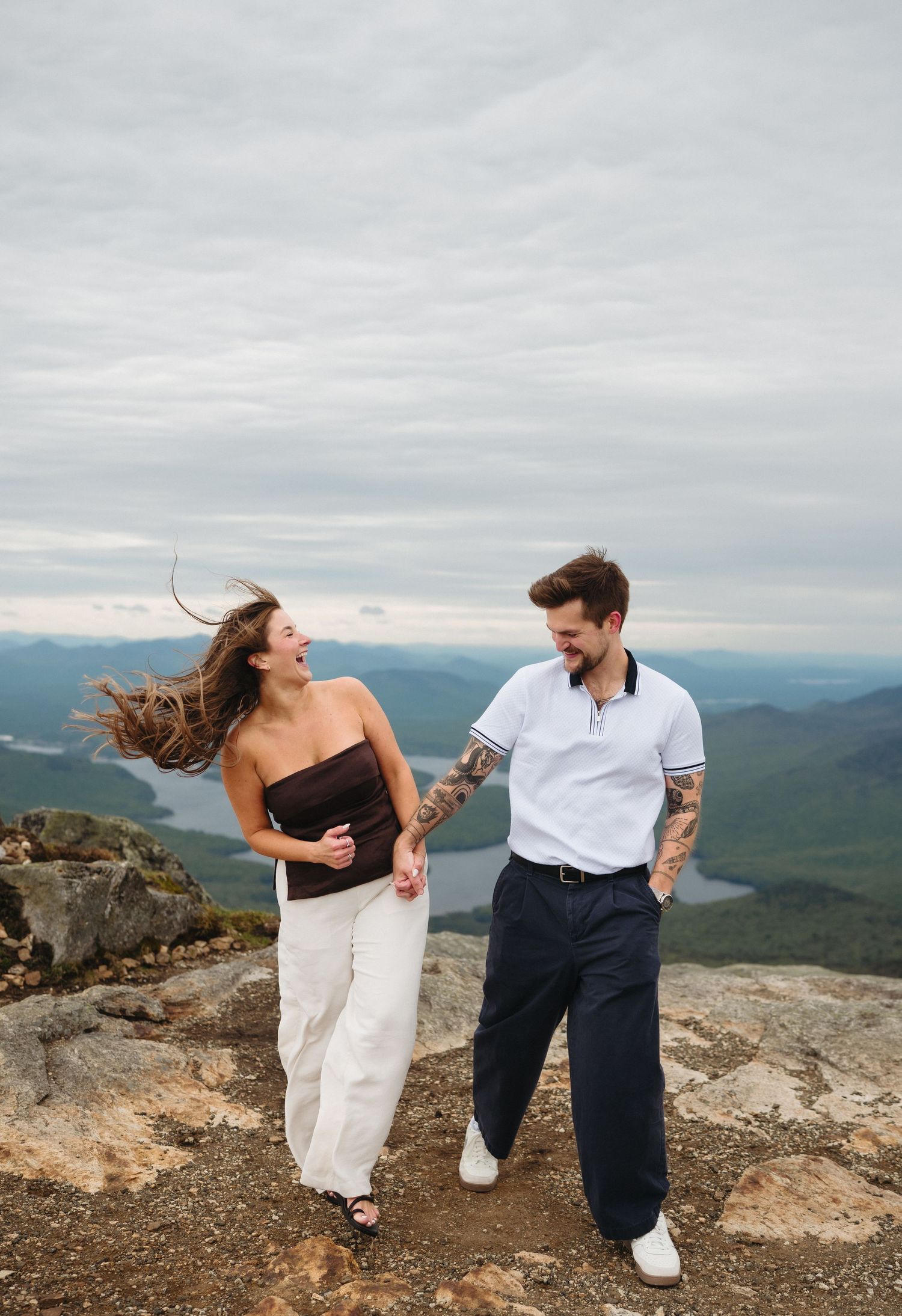 Couple laughing and holding hands on a mountain summit with scenic valley views on a cloudy day.
