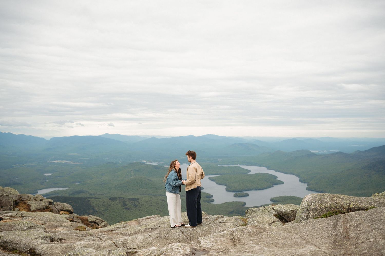 Couple embracing on rocky mountain summit with stunning aerial view of lake and green valley below on cloudy day.