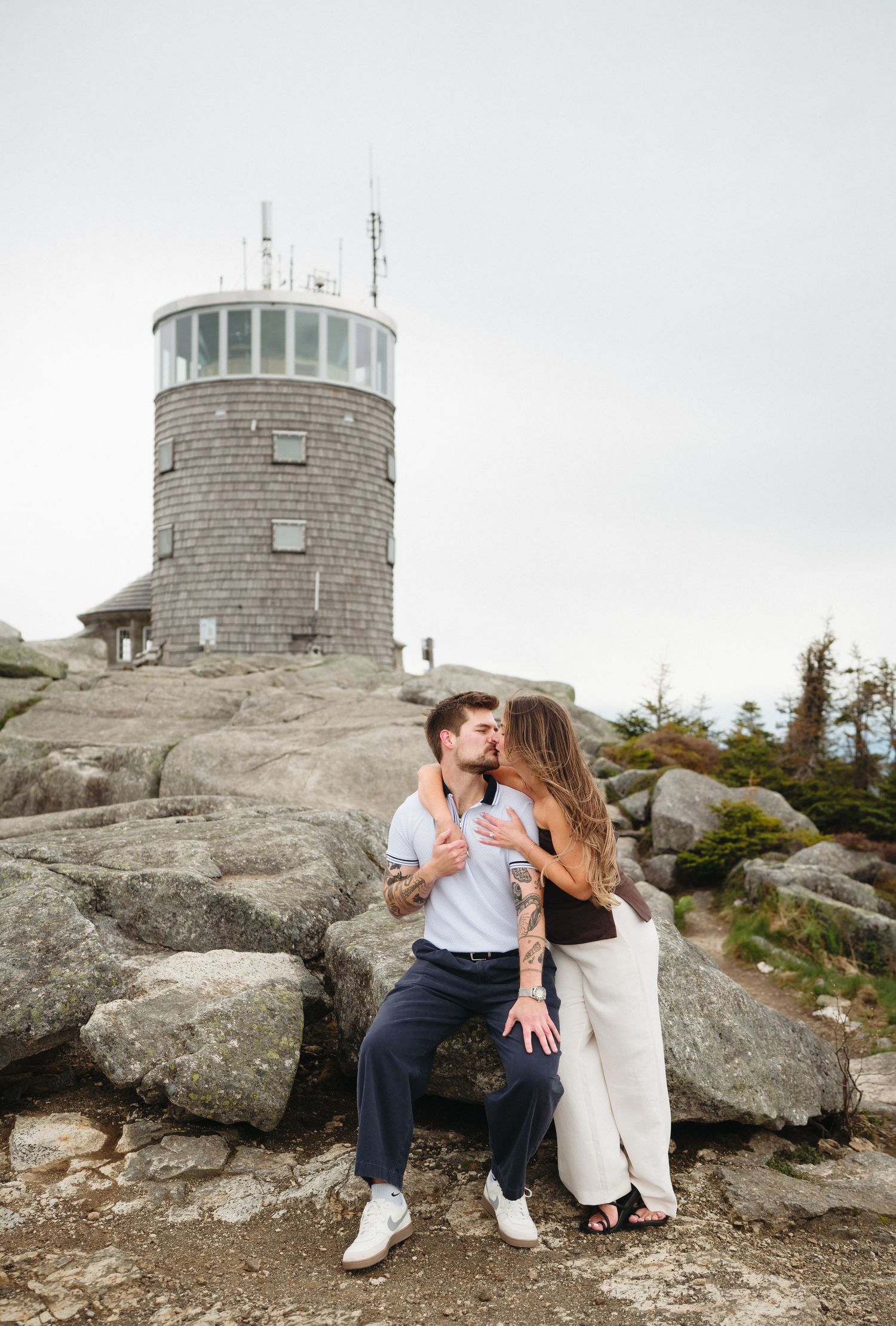 Couple sharing a romantic kiss on rocky terrain with a stone observation tower in the background on a cloudy day.