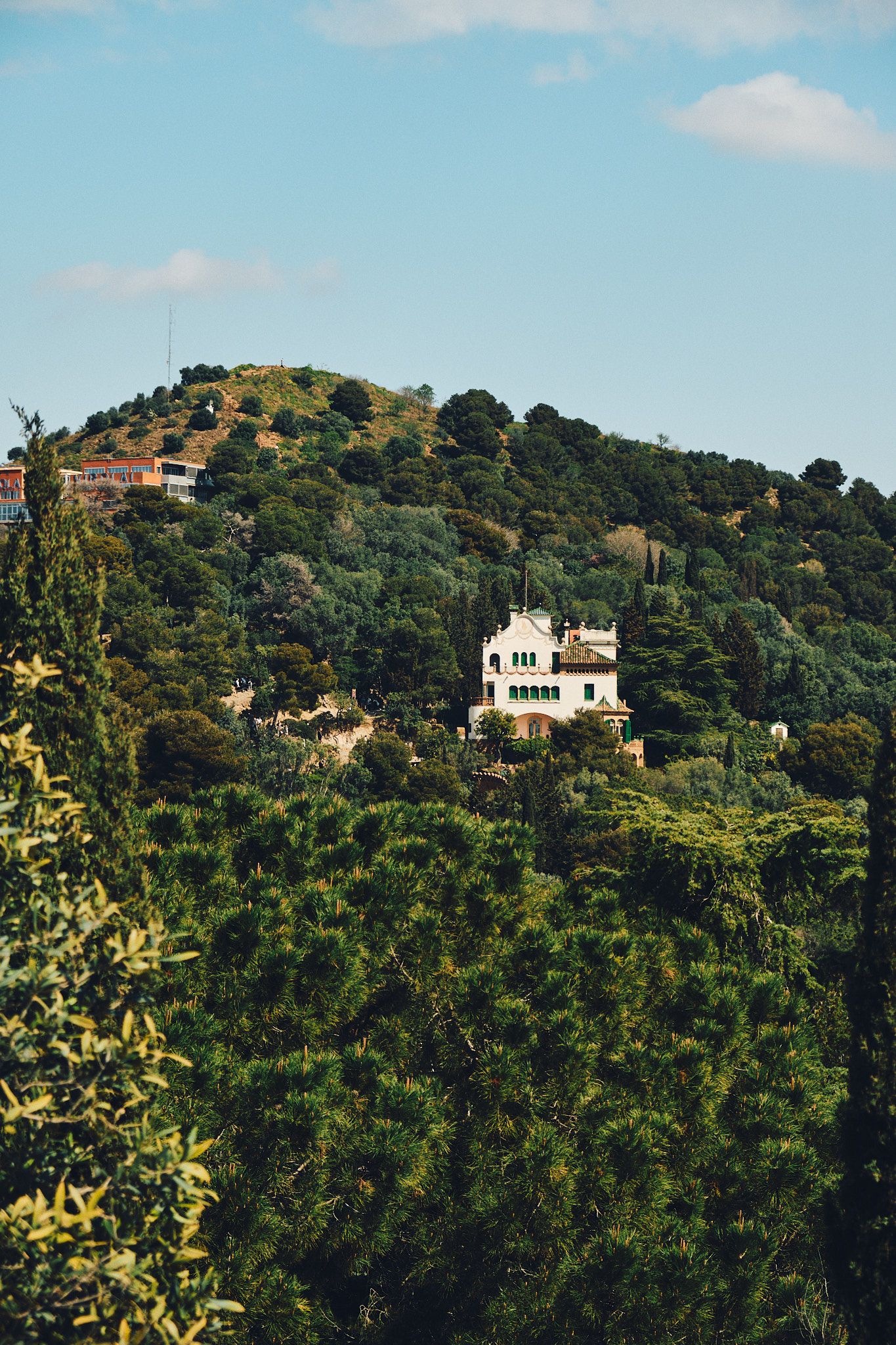 A white villa nestled among lush green trees on a hillside under a partly cloudy blue sky.