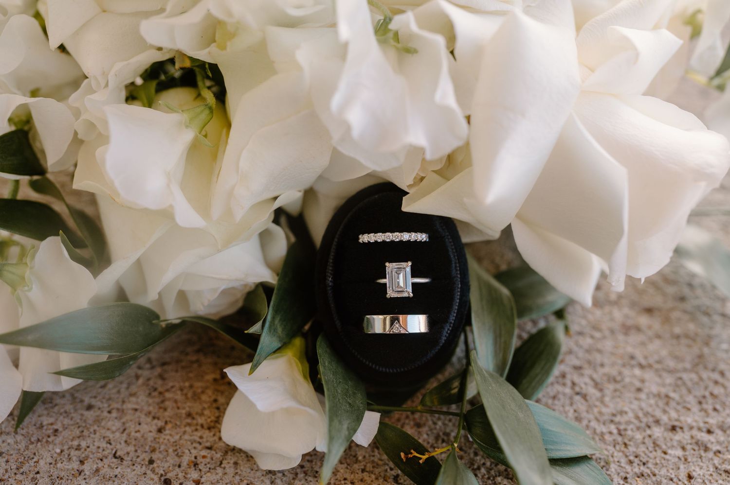 Elegant wedding rings displayed in a black velvet box surrounded by white roses and greenery for a bridal flat lay.