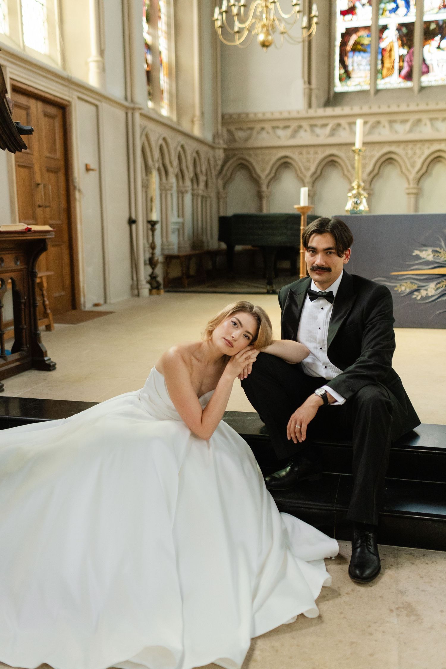 Elegant bride in white gown and groom in tuxedo sitting together in a grand Gothic church interior.