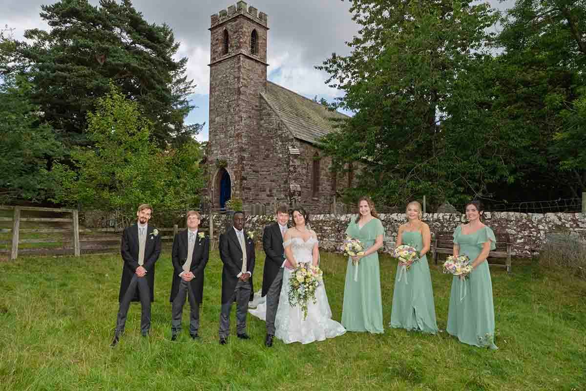 Wedding party posing outside a stone church, bride in white with bouquet, bridesmaids in sage green gowns, groomsmen in dark suits.