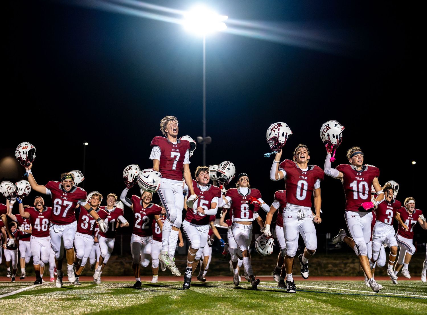 High school football team in maroon uniforms celebrating on field under bright stadium lights at night game.