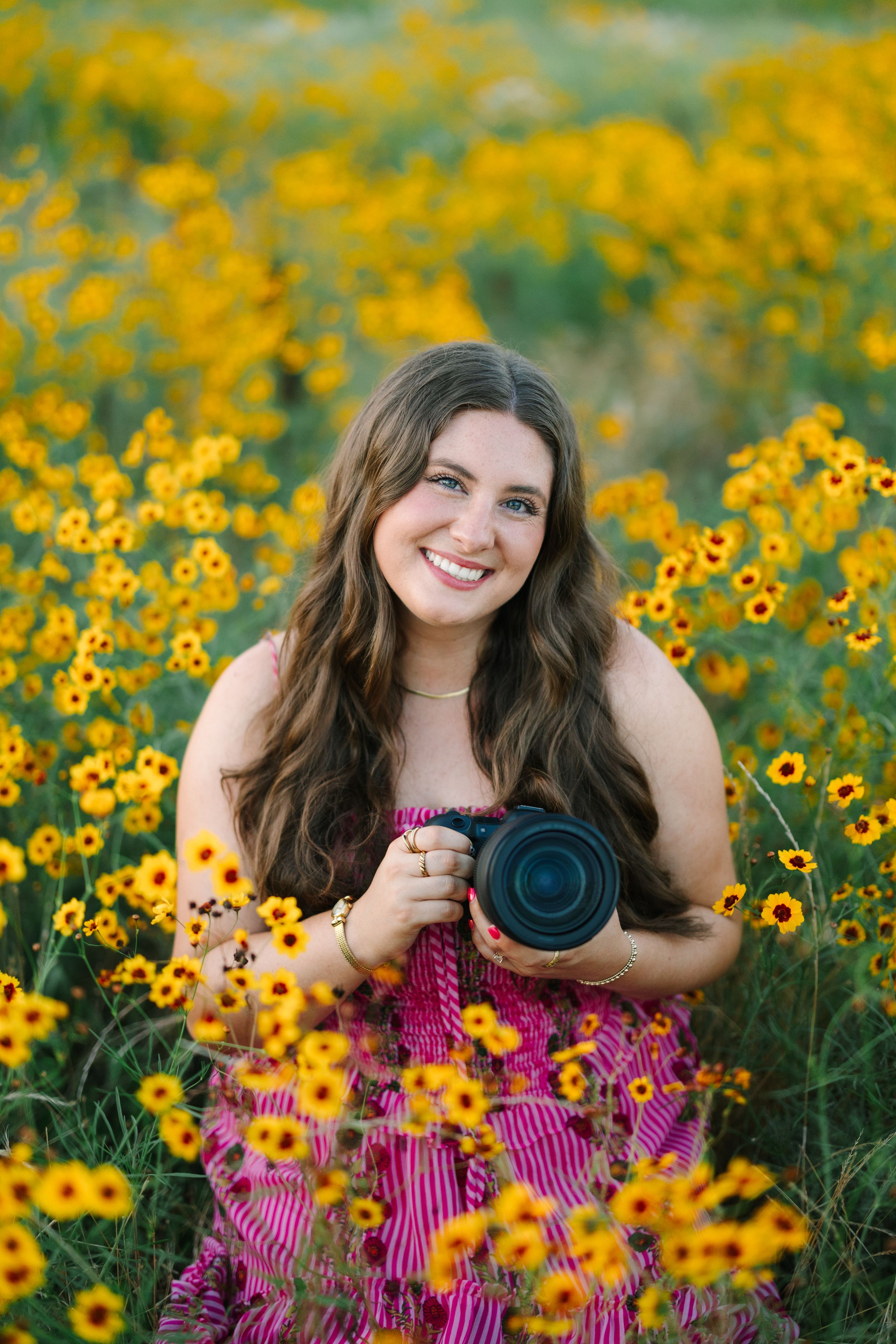 Young woman in pink dress holding a camera, smiling in a vibrant field of yellow wildflowers.