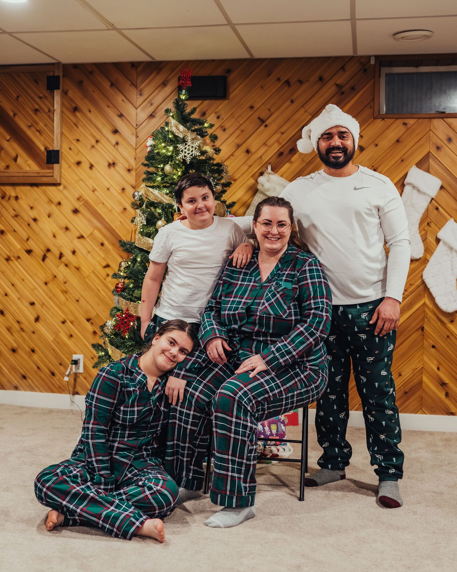 Family of four in matching plaid Christmas pajamas posing near a decorated tree with stockings on wood-paneled wall.