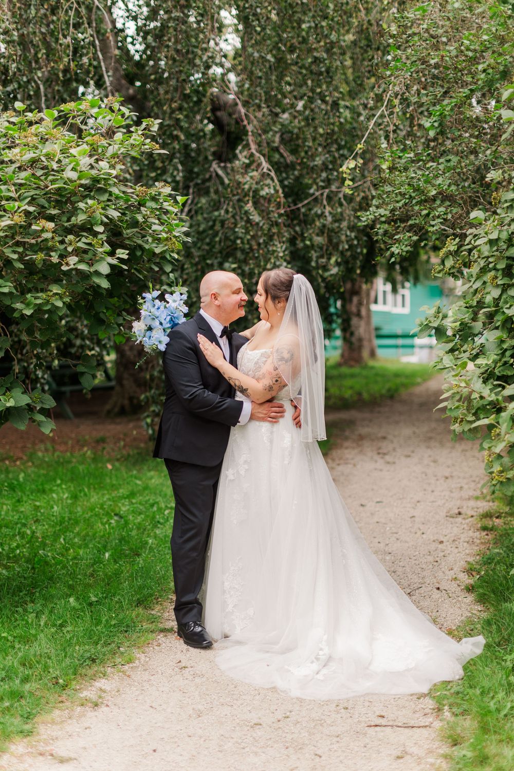 Bride and groom embrace on a garden path, she holds blue hydrangeas, wearing a white gown with veil, he wears a black suit.