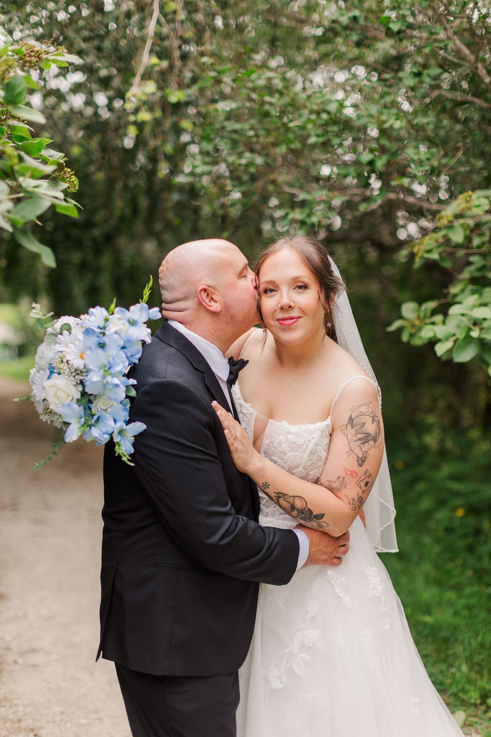 Groom kisses bride on cheek as she smiles, holding blue hydrangea bouquet, surrounded by lush greenery.
