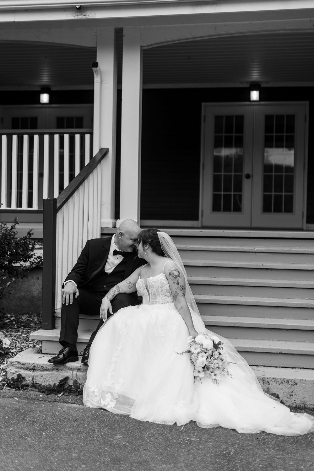 Black and white photo of bride and groom sitting on porch steps, sharing an intimate moment in wedding attire.