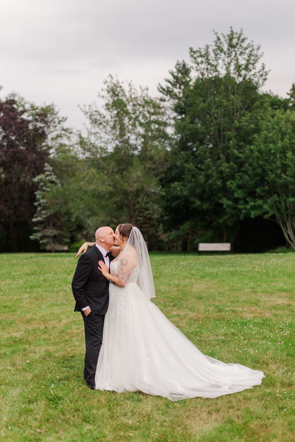Bride and groom share a romantic kiss on green lawn, wedding veil flowing, trees in background.