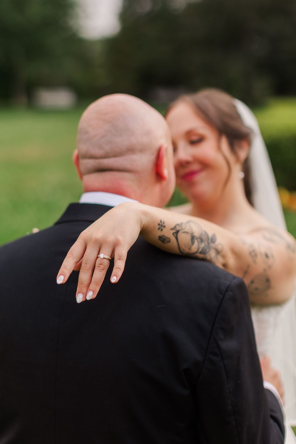 Bride with tattooed arm rests hand on grooms shoulder showing wedding ring, smiling softly in outdoor ceremony.