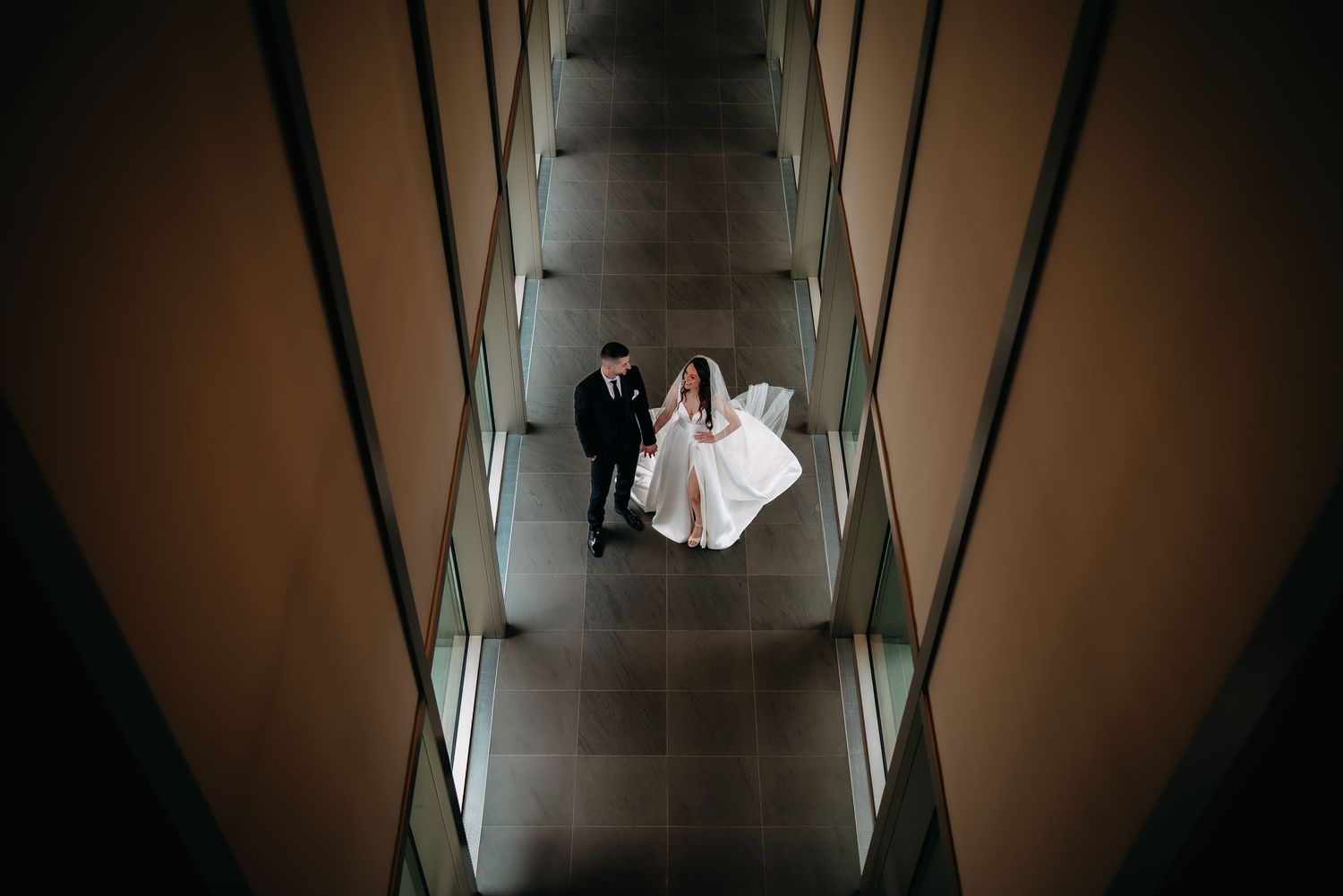 Aerial view of bride and groom walking through modern architectural corridor on wedding day.