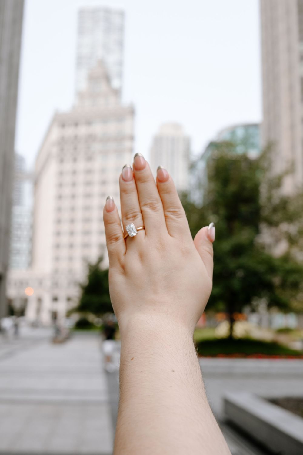 A hand with manicured nails displaying a stunning diamond engagement ring against a city skyline backdrop.