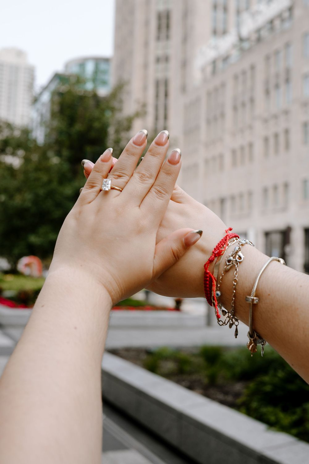 Close-up of two hands raised showing an engagement ring and charm bracelets against an urban backdrop.