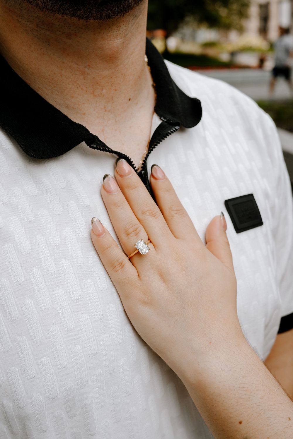 Close-up of a woman's hand displaying a diamond engagement ring on a gold band, resting on a man's white polo shirt.