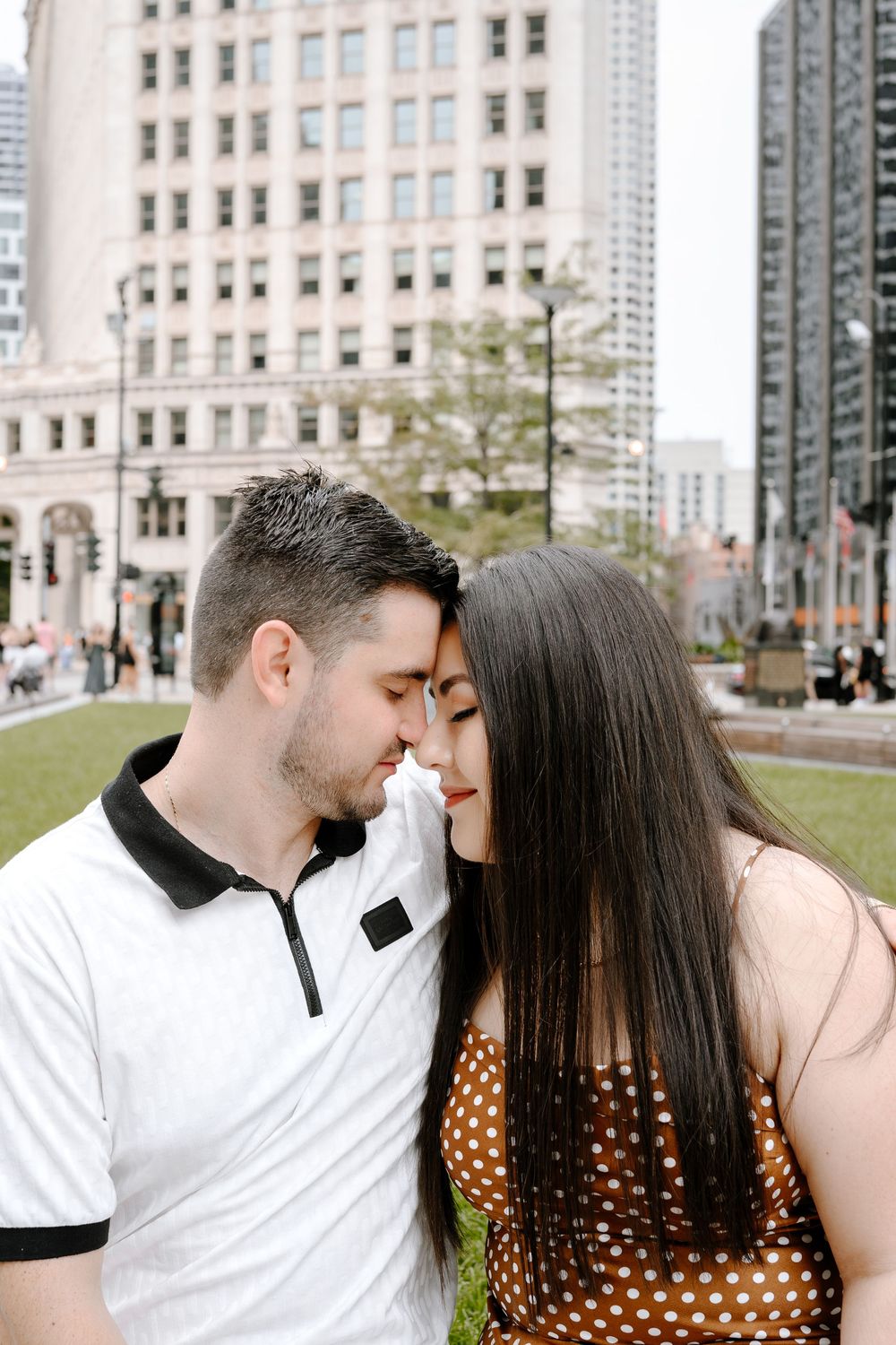 A couple shares a romantic nose-to-nose moment outdoors with city skyscrapers in the background.