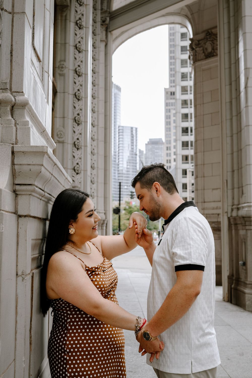 Couple sharing a romantic moment under an ornate stone arch in a city, man kissing womans hand while she smiles.