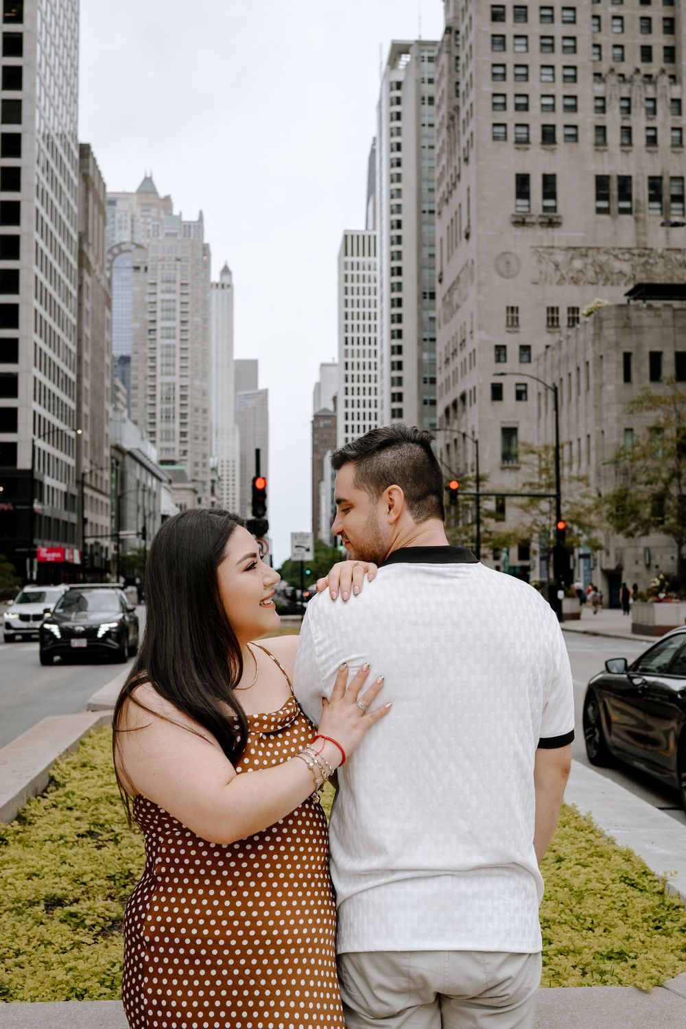 Couple embracing on a city street surrounded by tall buildings, woman in polka dot dress smiling at man in white shirt.