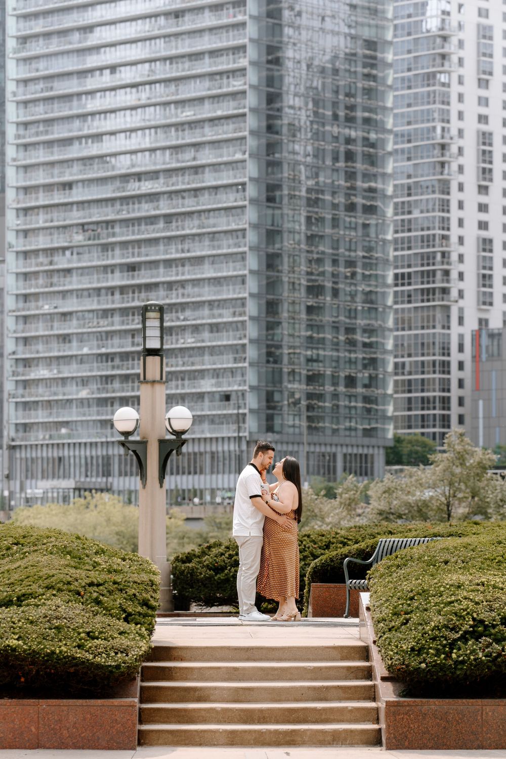 Couple embracing on steps in an urban park with modern glass skyscrapers in the background.