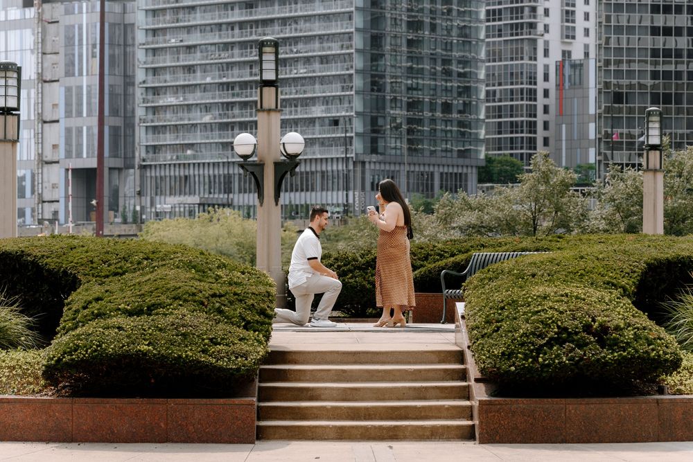 Man proposes on one knee to woman in urban park with modern skyscrapers in background.