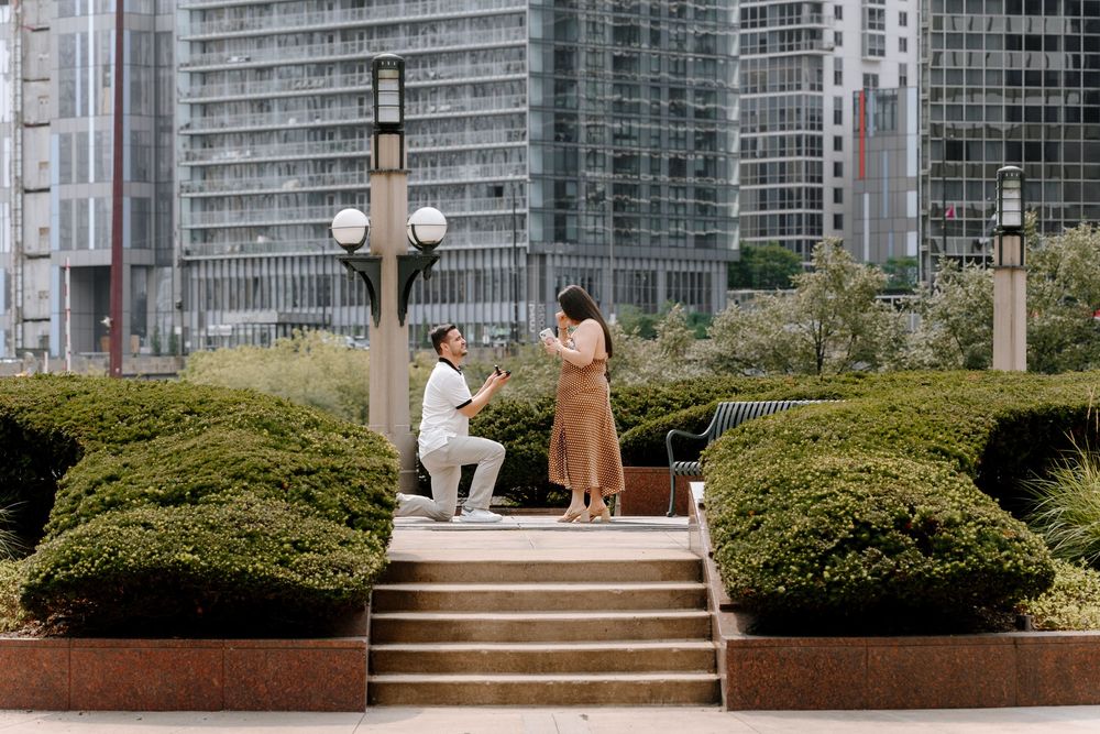 Man proposes to woman on one knee in urban garden with city skyscrapers in background.
