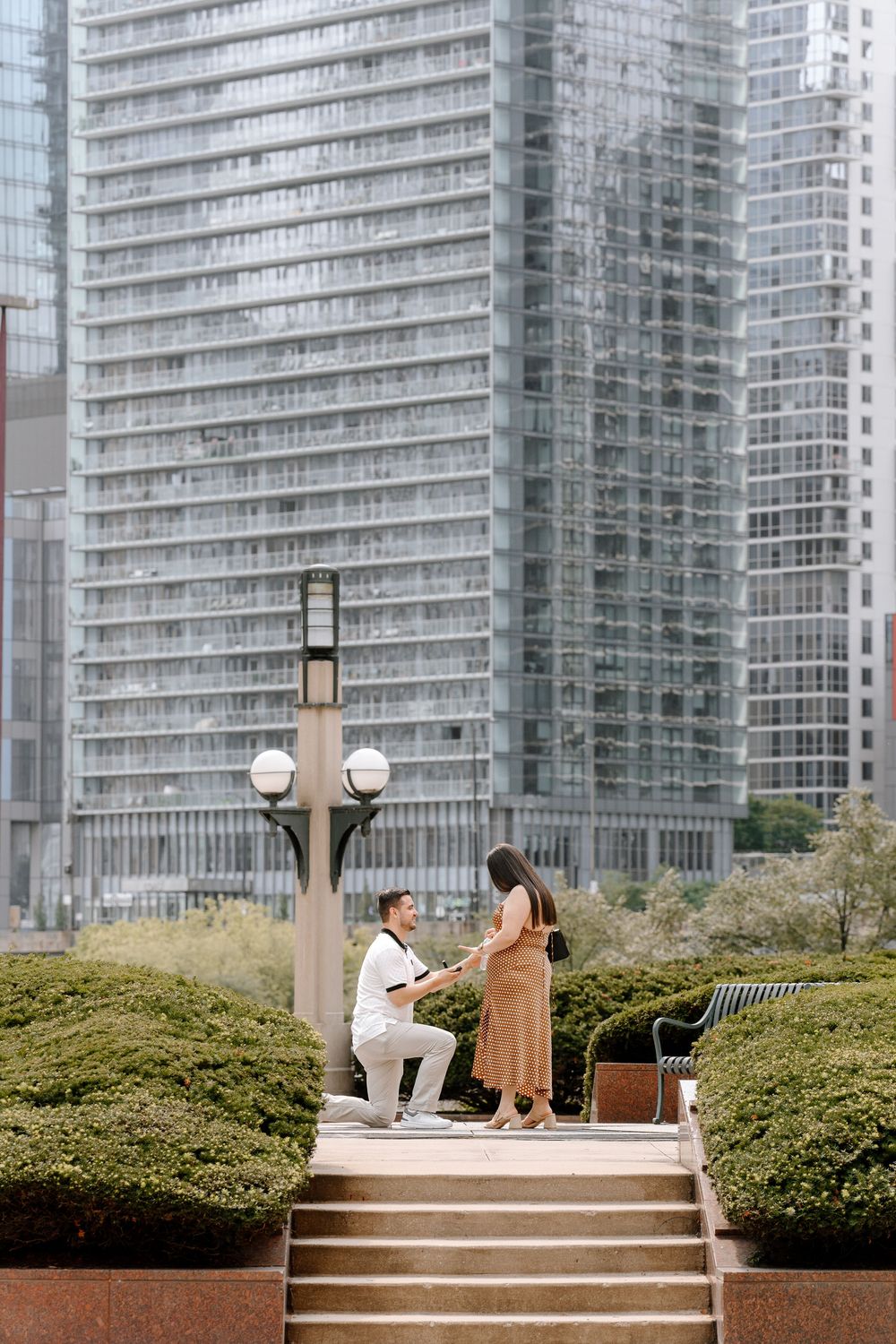 Man proposing on one knee to woman in tan dress near city skyscrapers and lamp post in urban park.