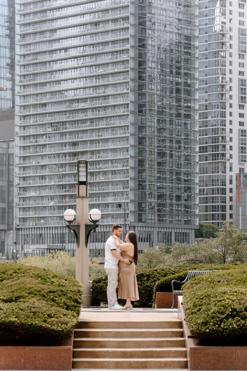 Couple embracing in an urban park with modern glass skyscrapers and lush green hedges in the background.