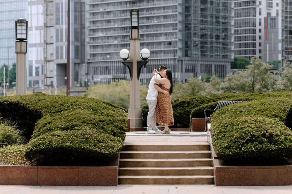 Couple embracing and kissing near a lamp post in an urban park with lush hedges and modern buildings.