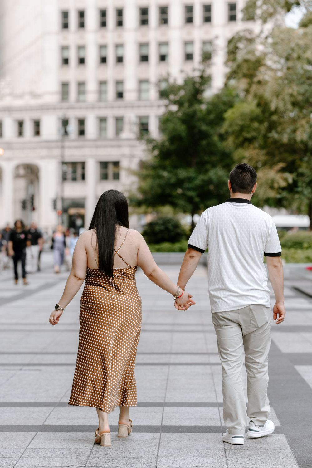 A couple holds hands while walking through an urban plaza, woman in brown polka dot dress, man in white polo and khaki pants.