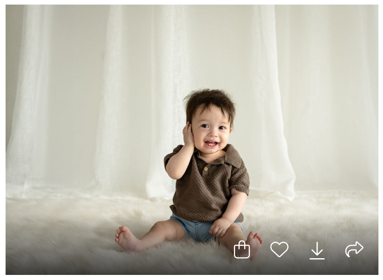 Adorable baby boy sitting on fluffy white rug, wearing brown top and denim shorts, posing cutely against white curtain backdrop.
