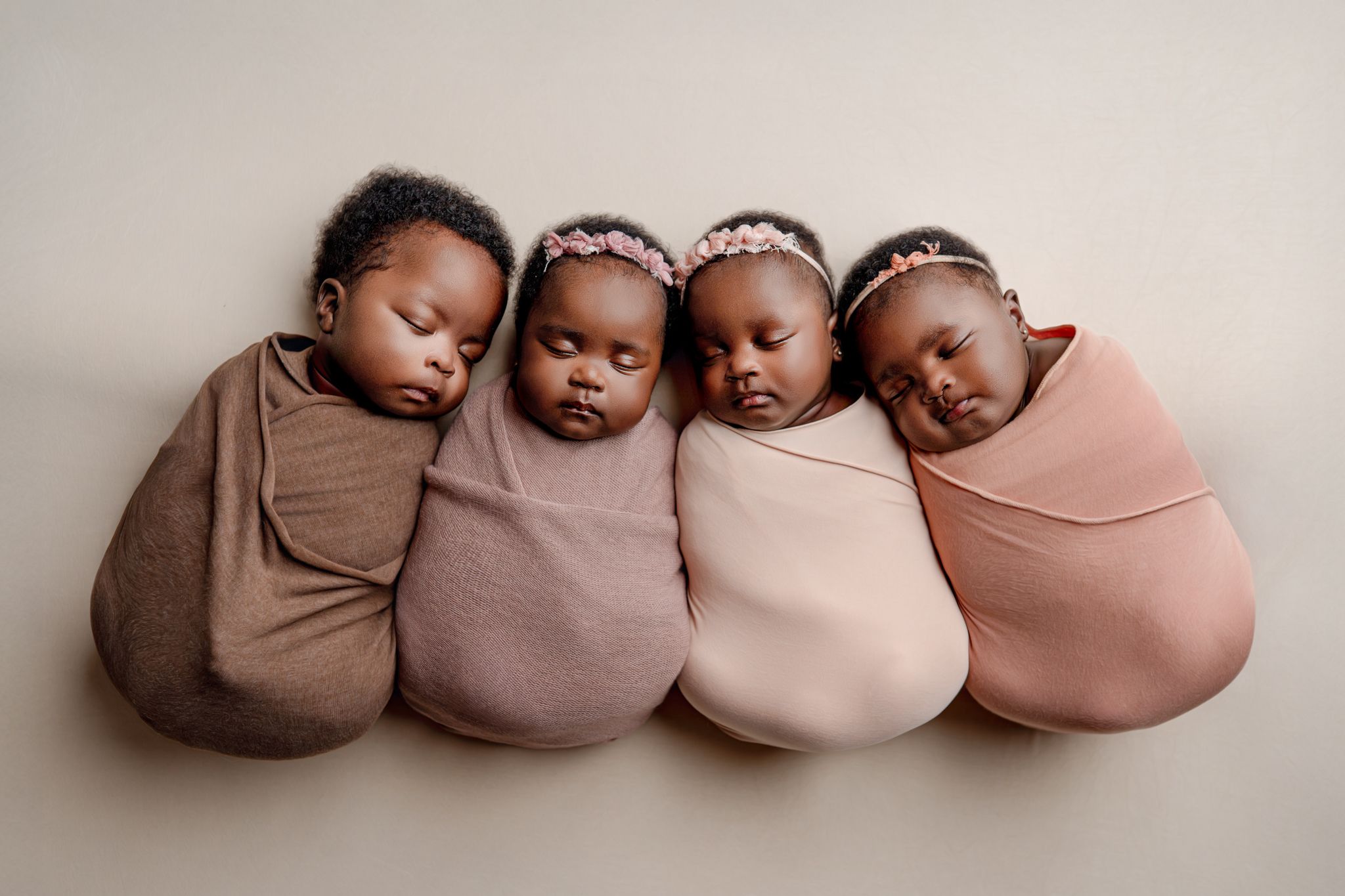 Four sleeping newborn babies swaddled in pink and brown wraps, lined up together in a peaceful studio portrait.