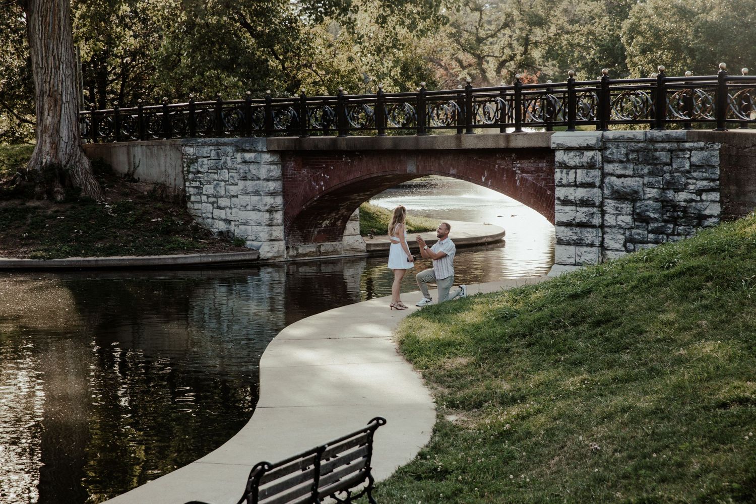 Couple holding hands near a stone arch bridge over a calm pond in a lush green park.