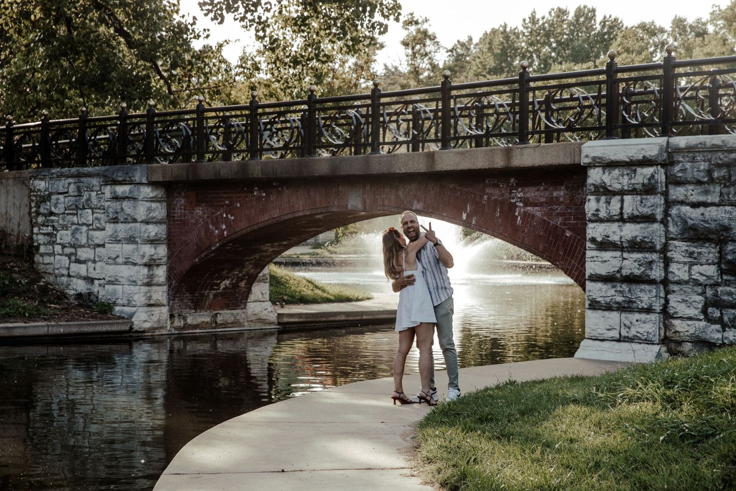 Couple kissing under a stone arch bridge by a calm river in a lush green park setting.