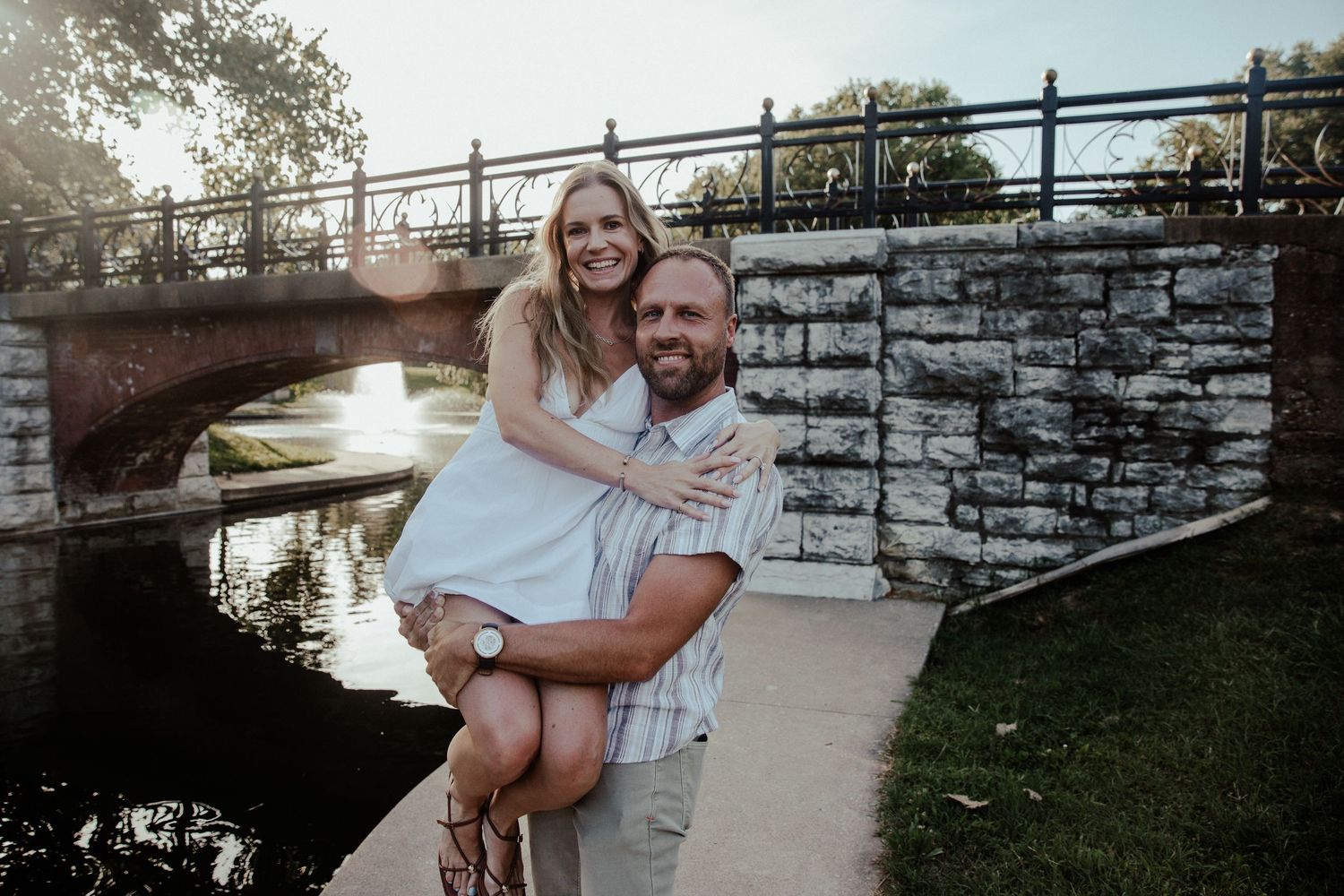 A couple poses happily by a stone bridge over a canal, the man carrying the woman in his arms outdoors.