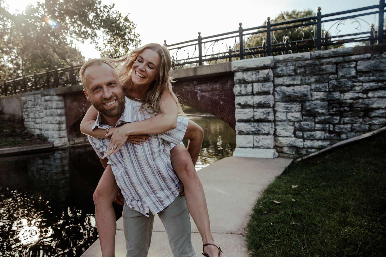 A smiling couple enjoys a piggyback ride outdoors near a stone bridge and pond on a sunny day.