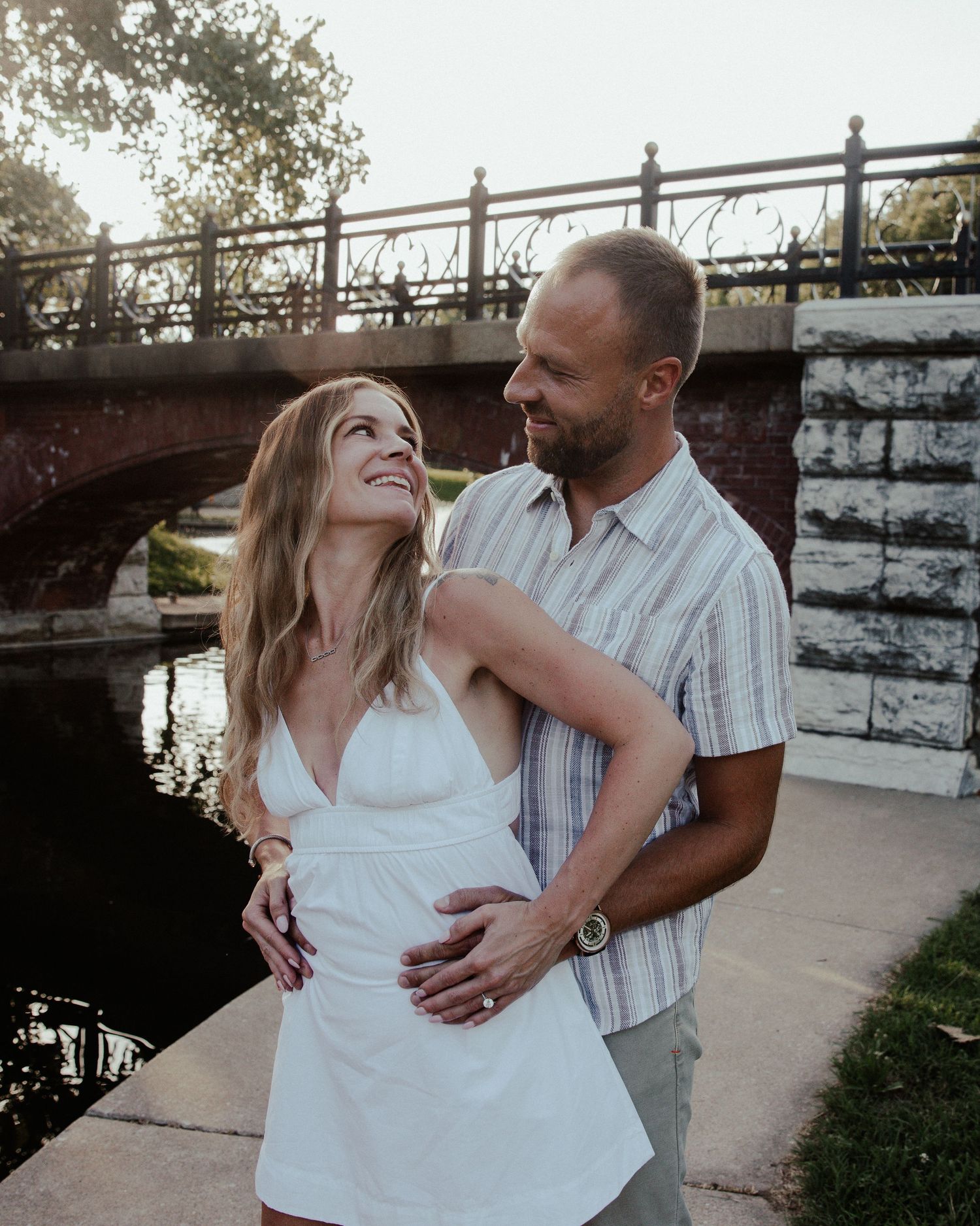 Couple embracing and smiling near a brick bridge over a canal, woman in white dress, man in plaid shirt.