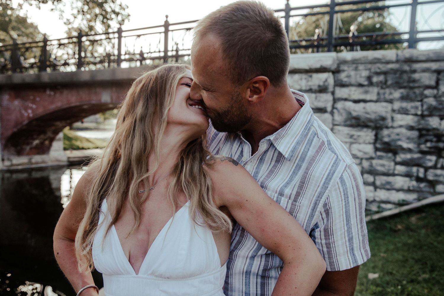 Couple sharing a romantic moment near a stone bridge, woman in white dress laughing as man leans in to kiss her.