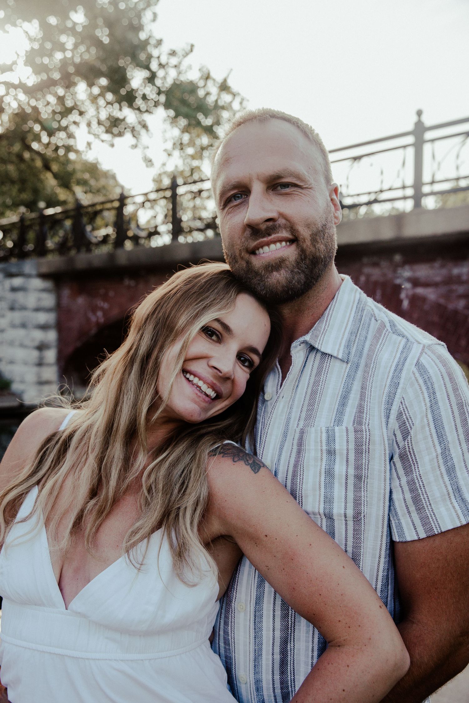 Smiling couple posing outdoors near a bridge, woman in white dress leaning on bearded man in striped shirt.