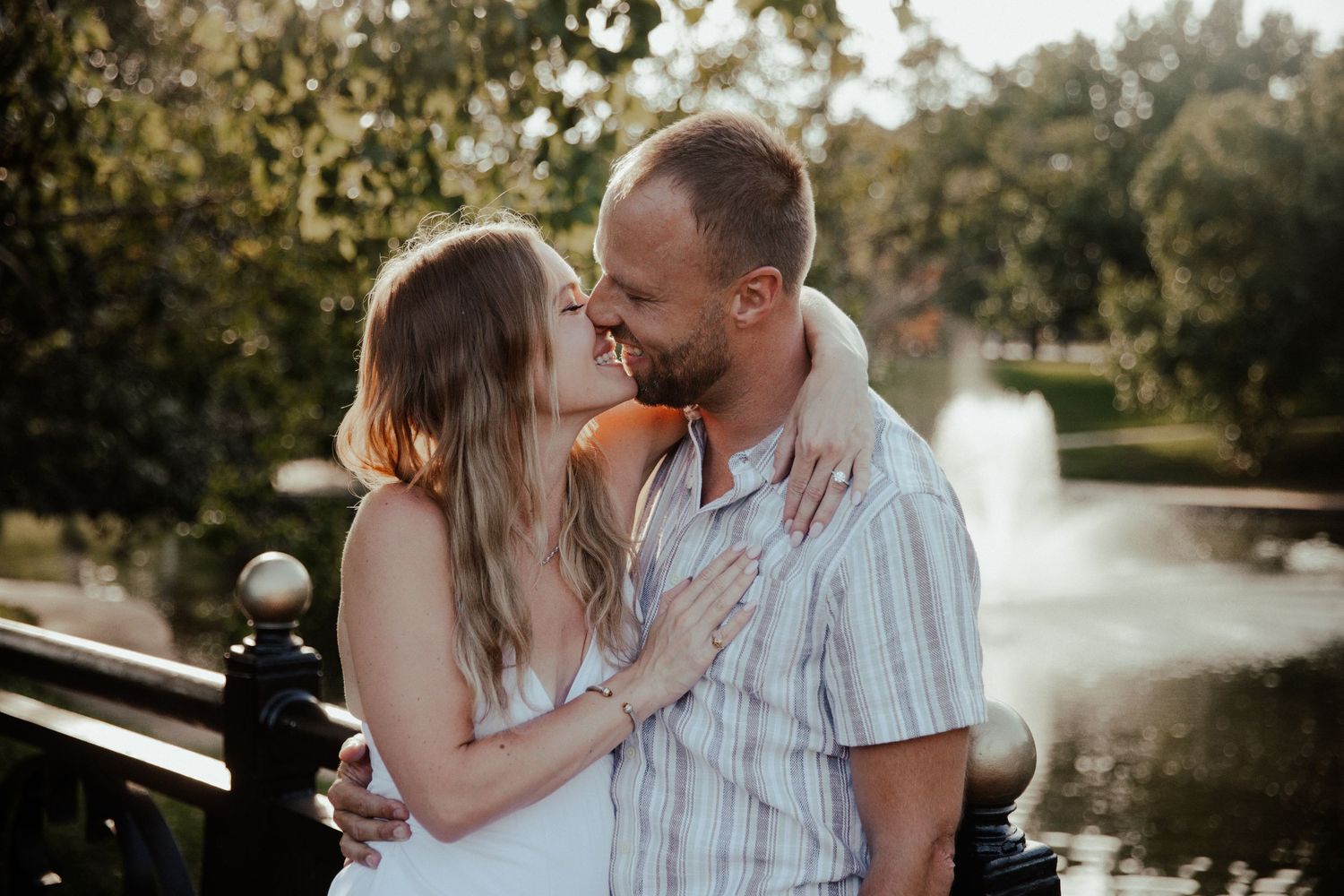 Couple sharing a romantic moment near a fountain in a lush green park on a sunny day.