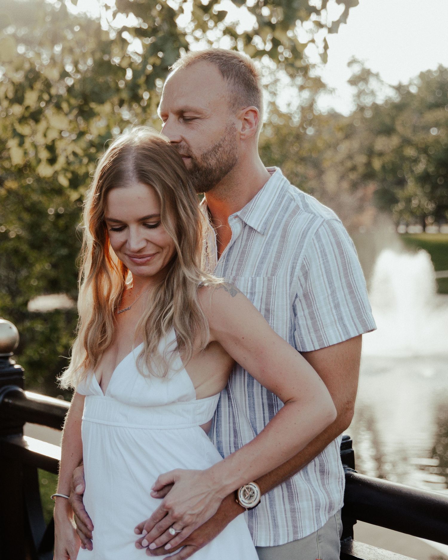 Couple embraces tenderly near a fountain in golden hour light, woman in white dress.