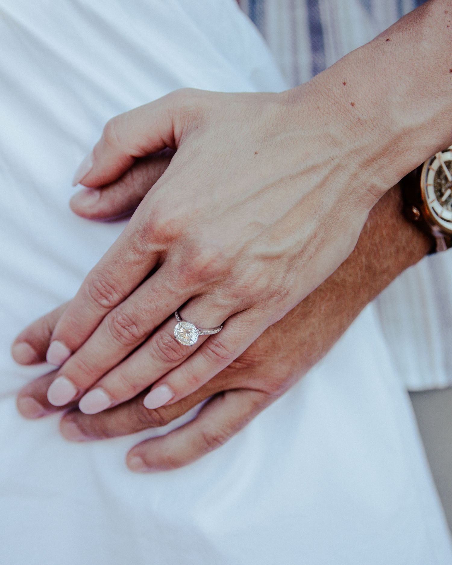 Close-up of a woman's hand wearing a stunning oval diamond engagement ring, resting on a man's hand wearing a gold watch.