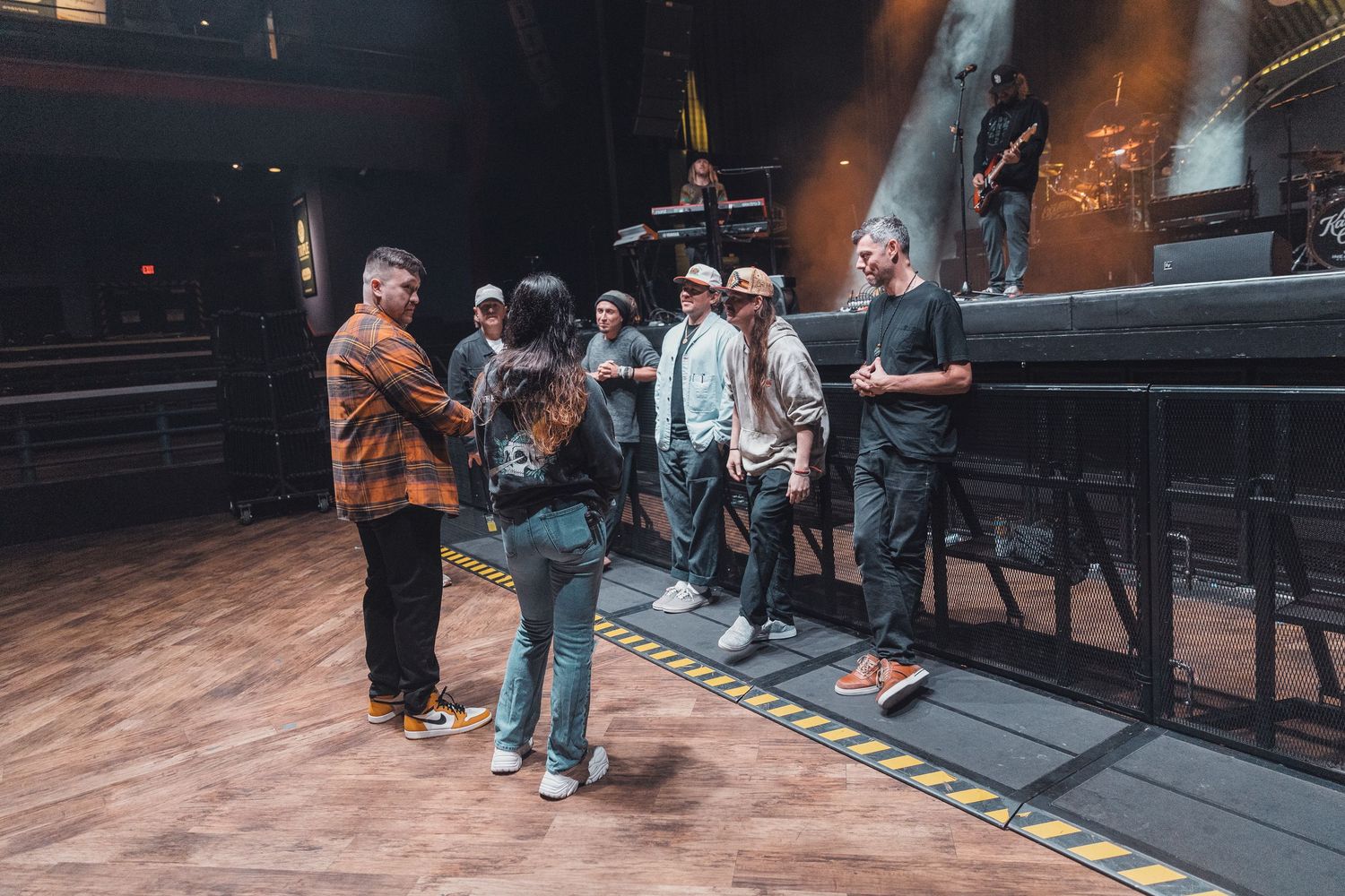 Group of people gathered on a wooden stage near concert equipment in a dimly lit venue.