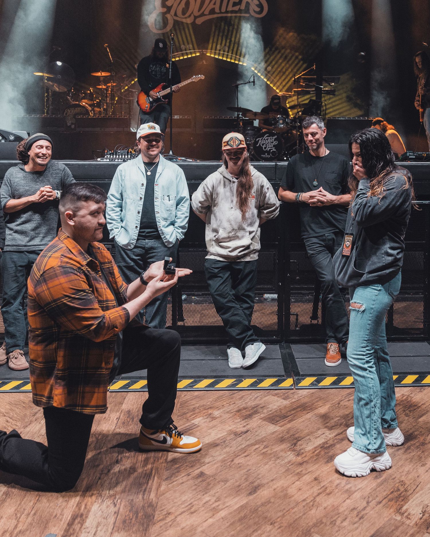 Man proposes on stage at a concert venue while bandmates watch and celebrate in the background.